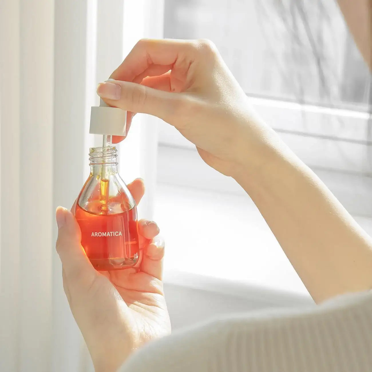 Hands holding AROMATICA skincare serum bottle with dropper, natural light, holistic beauty routine