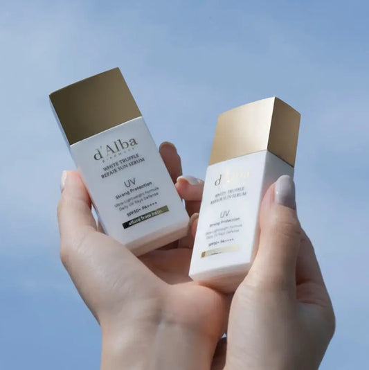 Hands holding d'Alba White Truffle Repair Sun Serum bottles with SPF50+ UV protection against blue sky